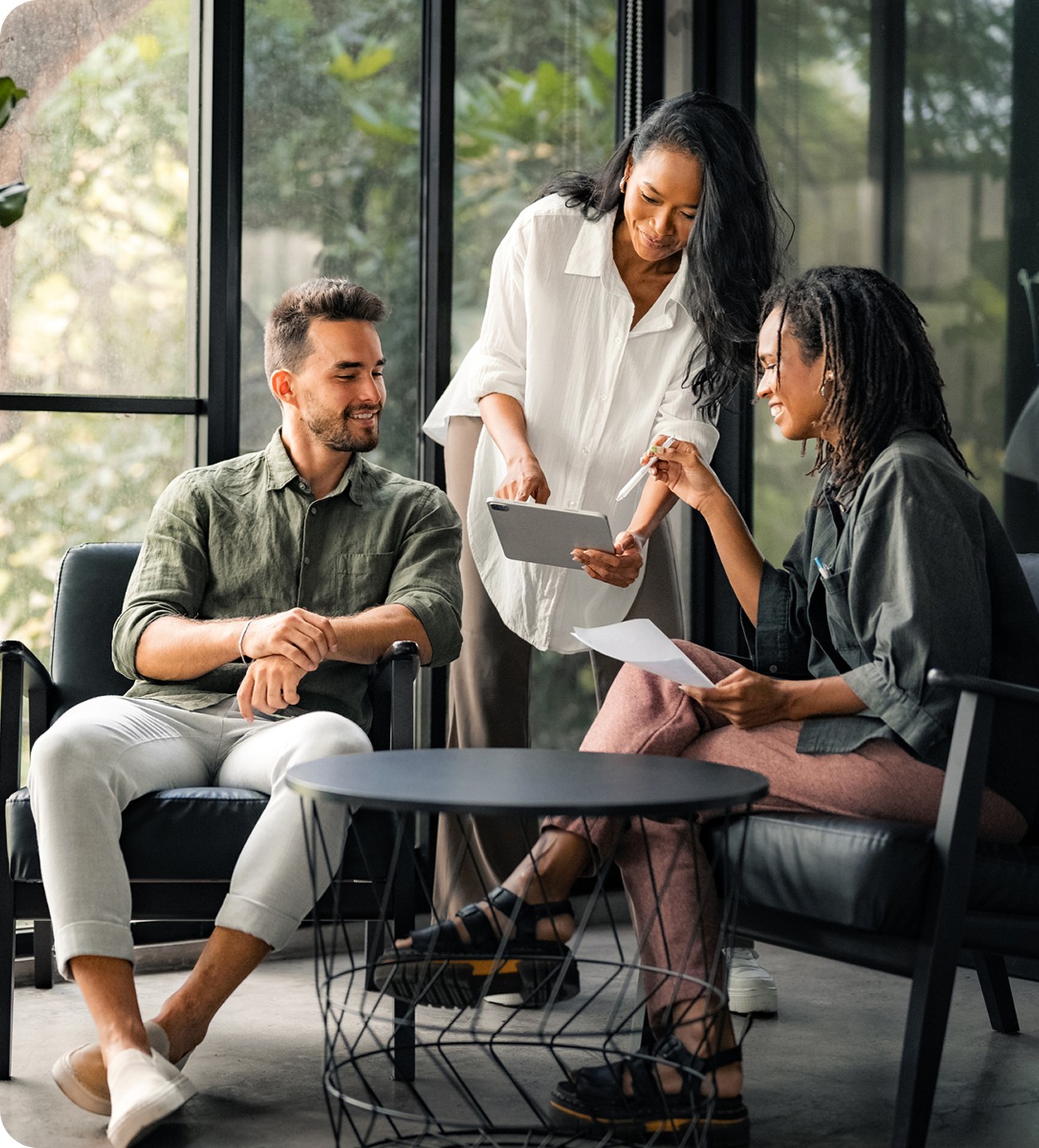A group of three colleagues collaborating happily in a modern office space, sitting around a small coffee table with a tablet and documents.