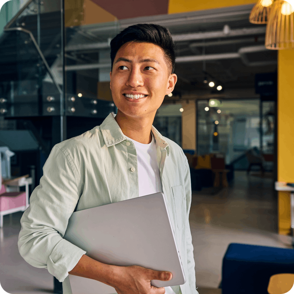 Front-facing view of a smiling young professional holding a laptop in a modern, brightly lit office environment. Front-facing view of a smiling young professional holding a laptop in a modern, brightly lit office environment.