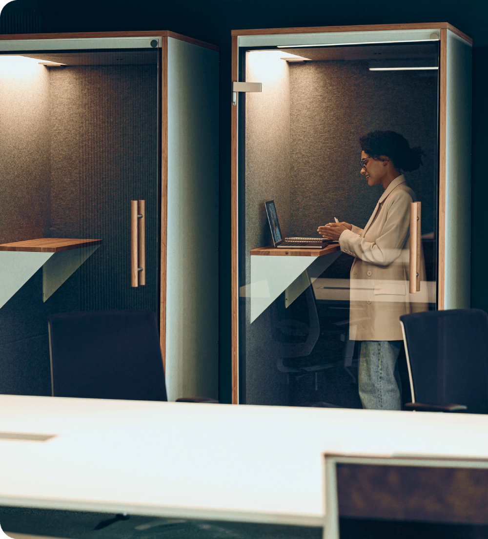 A workplace scene featuring two glass-walled phone booths, with a person sitting inside one booth working on a laptop.