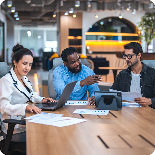 Three diverse corporate colleagues seated together around a modern conference desk working on laptops and tablets in a brightly lit office space