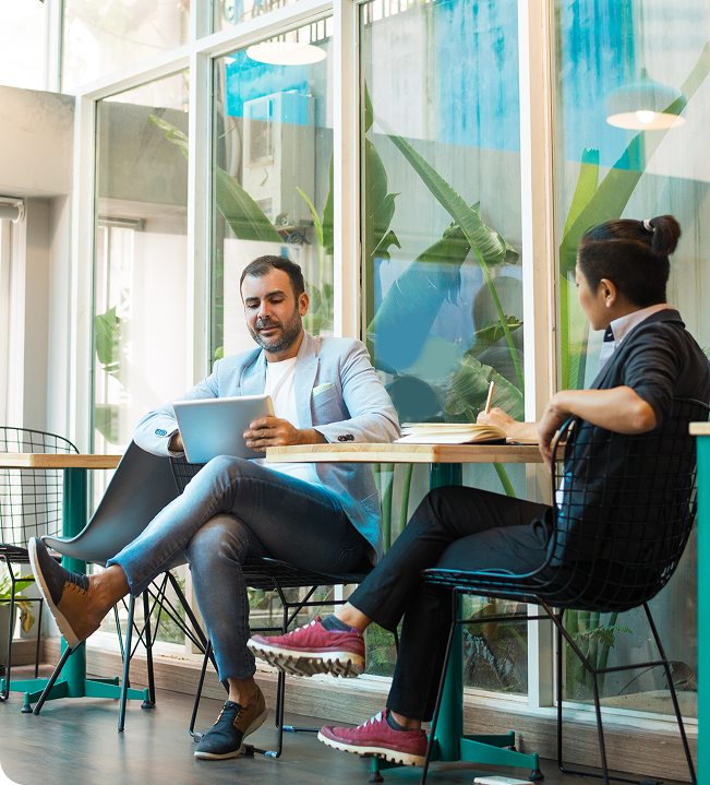 Two office colleagues sitting at a modern wooden desk, working together on a laptop in a brightly lit, contemporary workspace